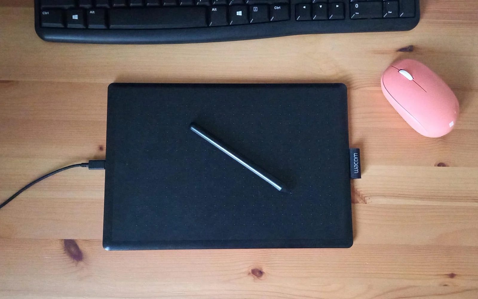 Svart tegnebrett med penn på et trebord, med tastatur og rosa mus ved siden av.