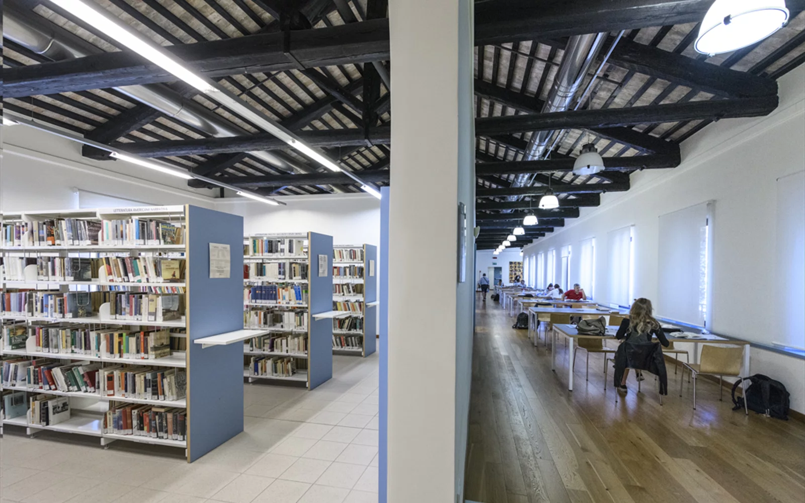 Bibliotek med bokhyller, mørke takbjelker og leseplasser langs store vinduer.