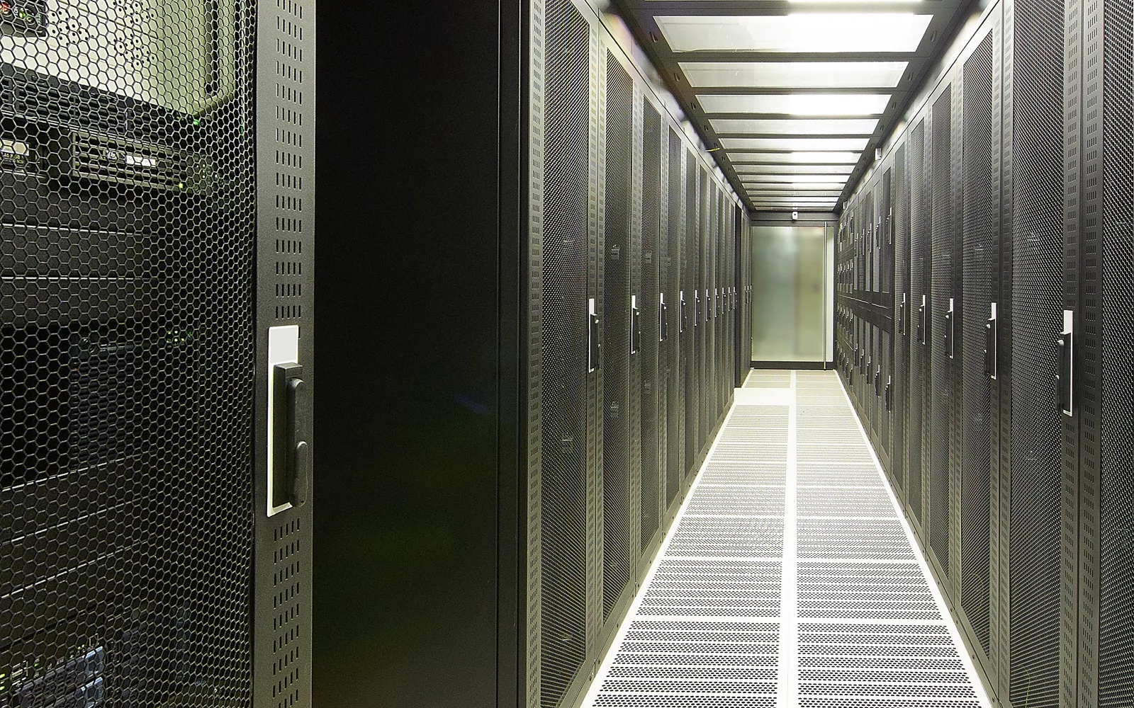 Smal gang mellom svarte serverrack, med ventilasjonsrister i gulvet.