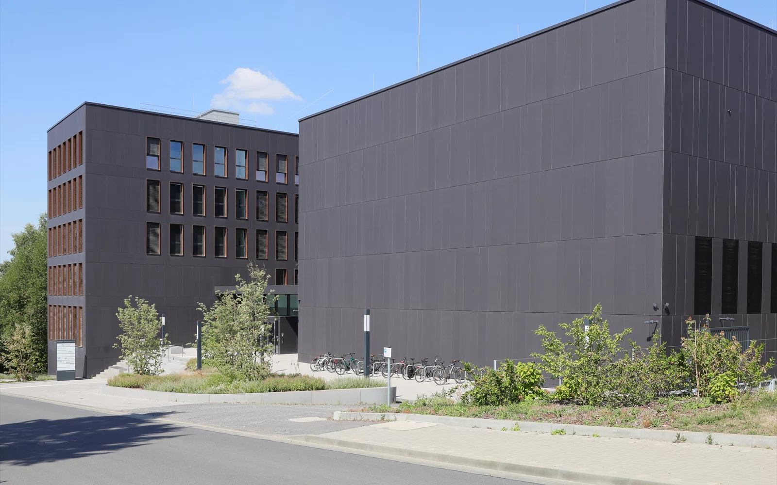 Moderne bygningskompleks med mørk sidevegg og beplantning foran.
