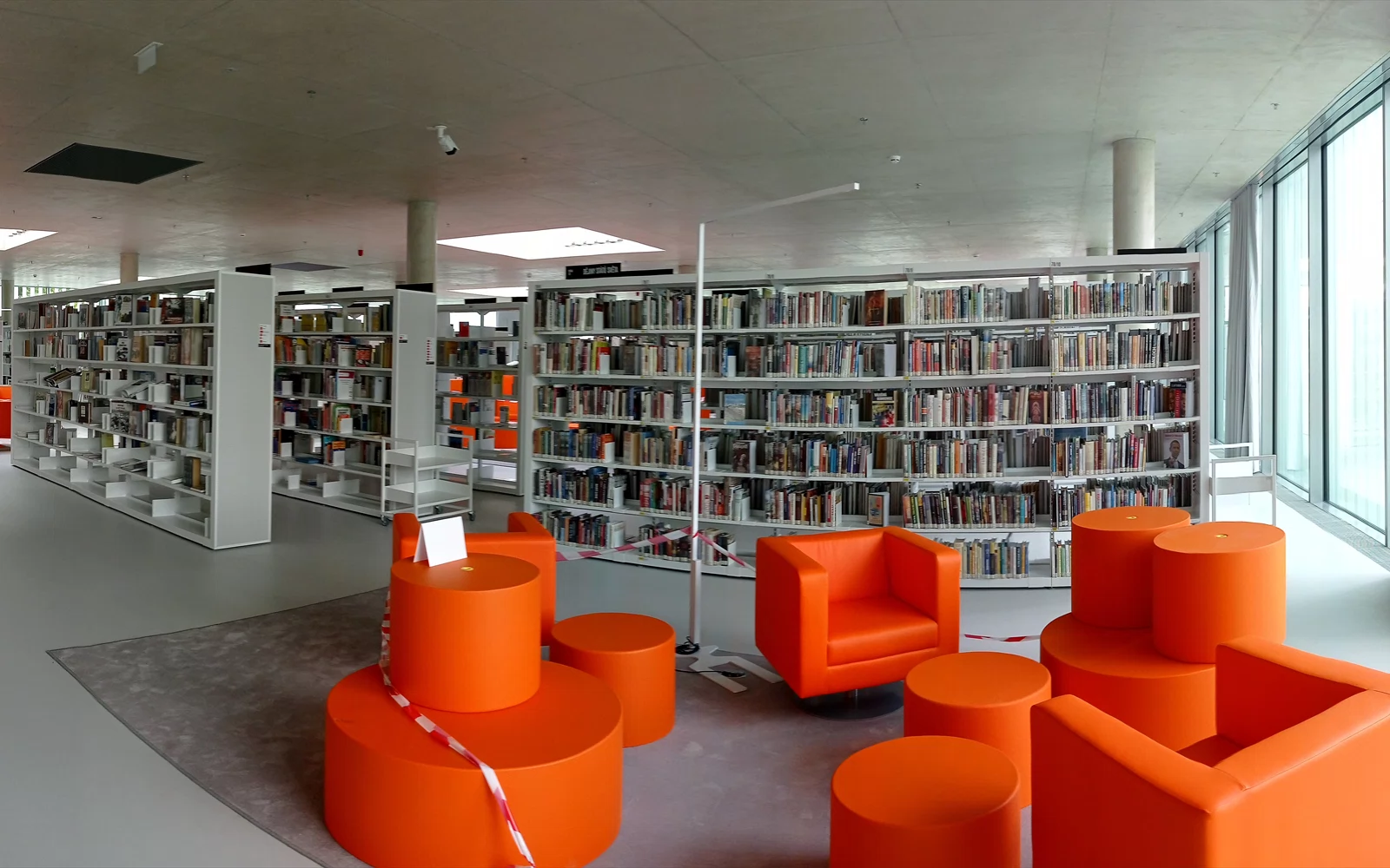 Moderne bibliotek med bokhyller, store vinduer og oransje lenestoler.