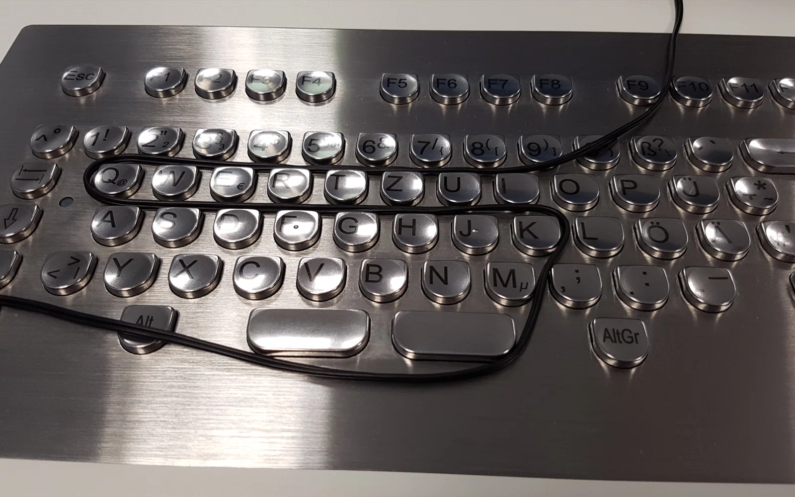 Tastatur i børstet metall med runde taster og en svart ledning.