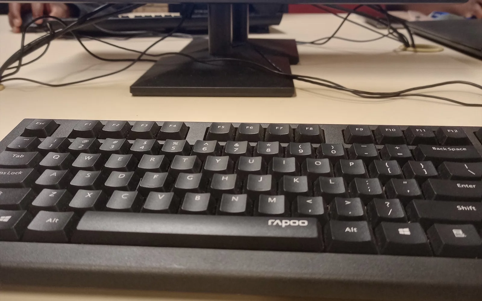 Svart tastatur på et skrivebord foran en skjermfot og flere kabler.